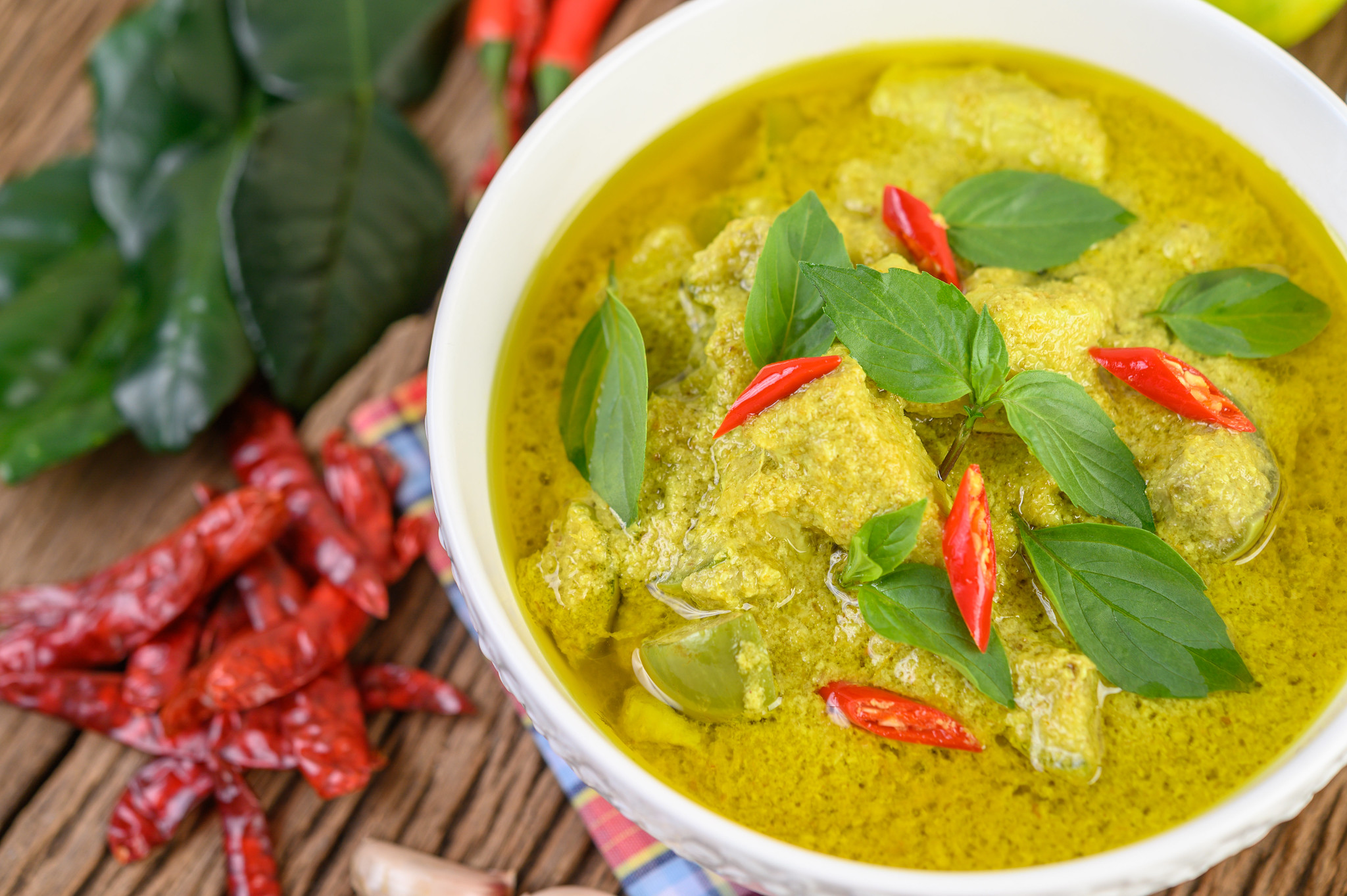 Vibrant green curry served in a bowl, garnished with fresh lime wedges, thinly sliced red onion, kaffir lime leaves, lemongrass, and garlic; a fragrant, colourful snapshot of Thai cooking